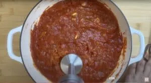 Blending homemade tomato sauce in a white pot with an immersion blender on a wooden table.